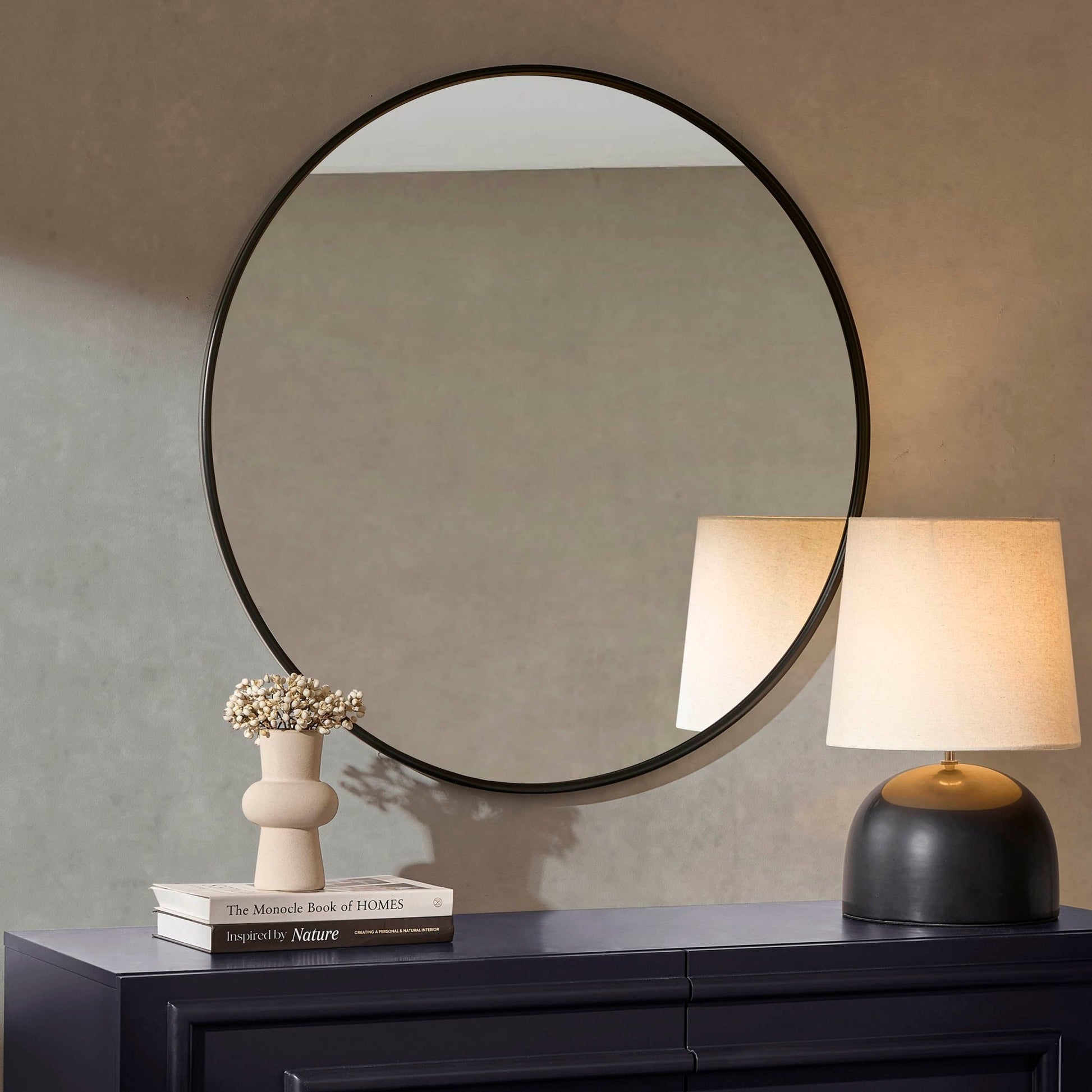 Modern round hallway mirror with black frame, styled above a navy console with stacked books, a ceramic vase, and soft-glow table lamp.